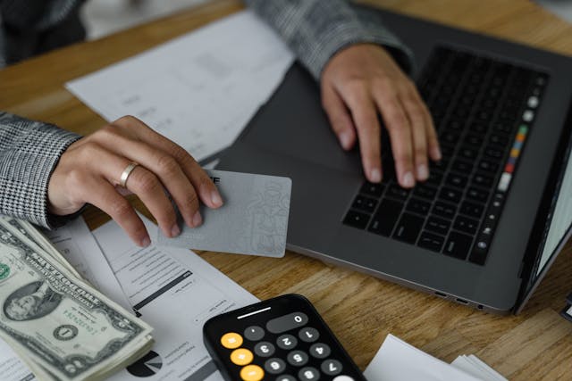 Person using a laptop while holding a bank card with money and a calculator sitting on the table