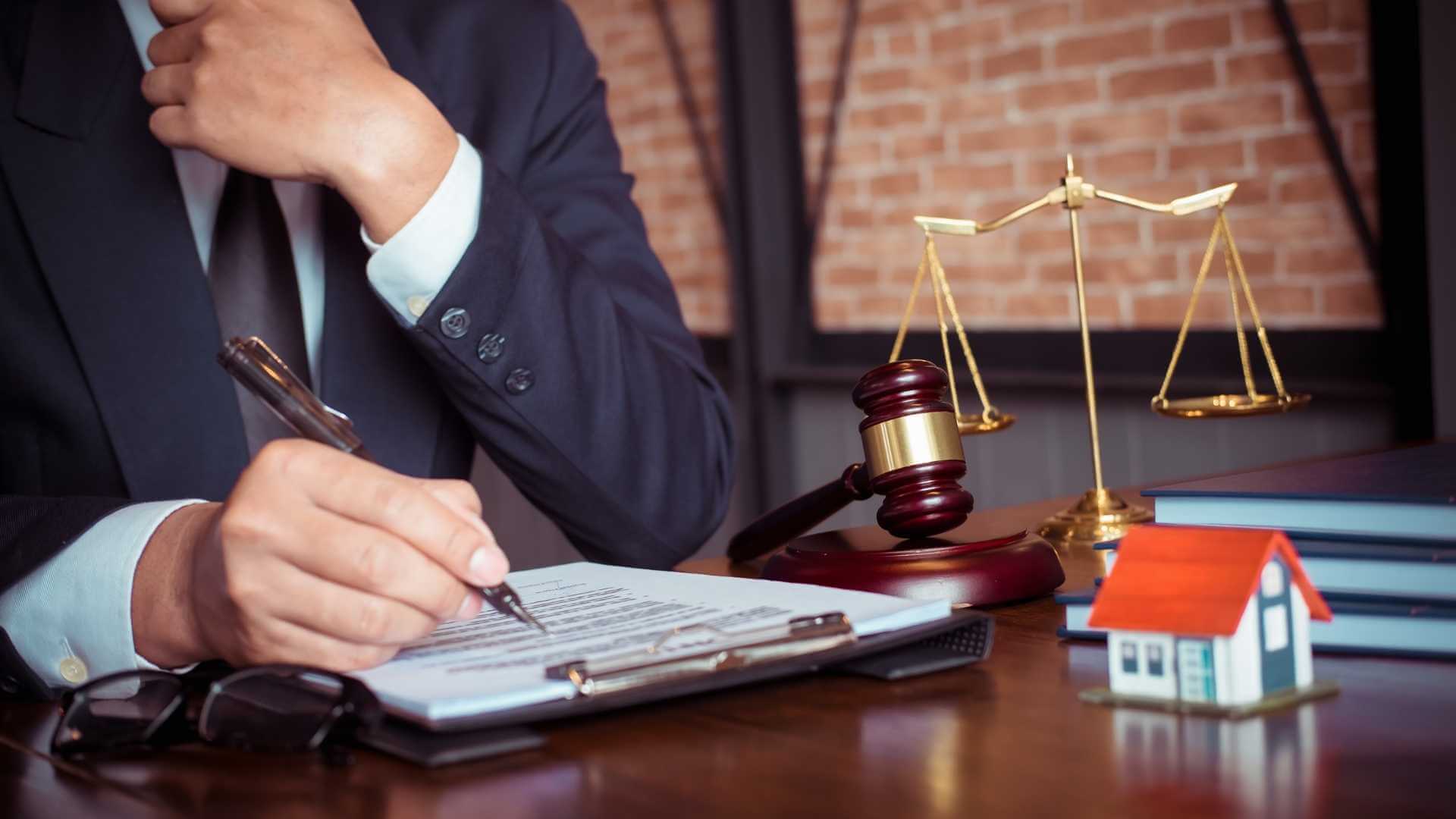 person sitting at a desk reviewing a contract with a gavel, house model, and balance scale next to it